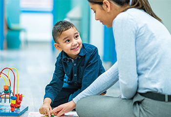 An SLP and a young boy sit on the floor next to a toy and point to a worksheet.